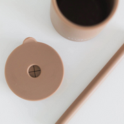A close-up of the cocoa-colored silicone sippy cup with its lid removed, showing the built-in straw hole and spill-resistant cross slit. The straw is placed beside the cup, highlighting its durable, easy-to-clean design.