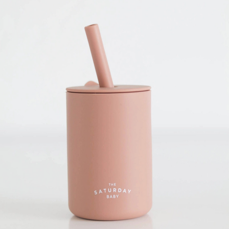 A coral-colored silicone straw cup from The Saturday Baby, featuring a sleek, minimalist design with a matching lid and straw. The cup is placed against a clean white background, highlighting its modern and functional aesthetic.