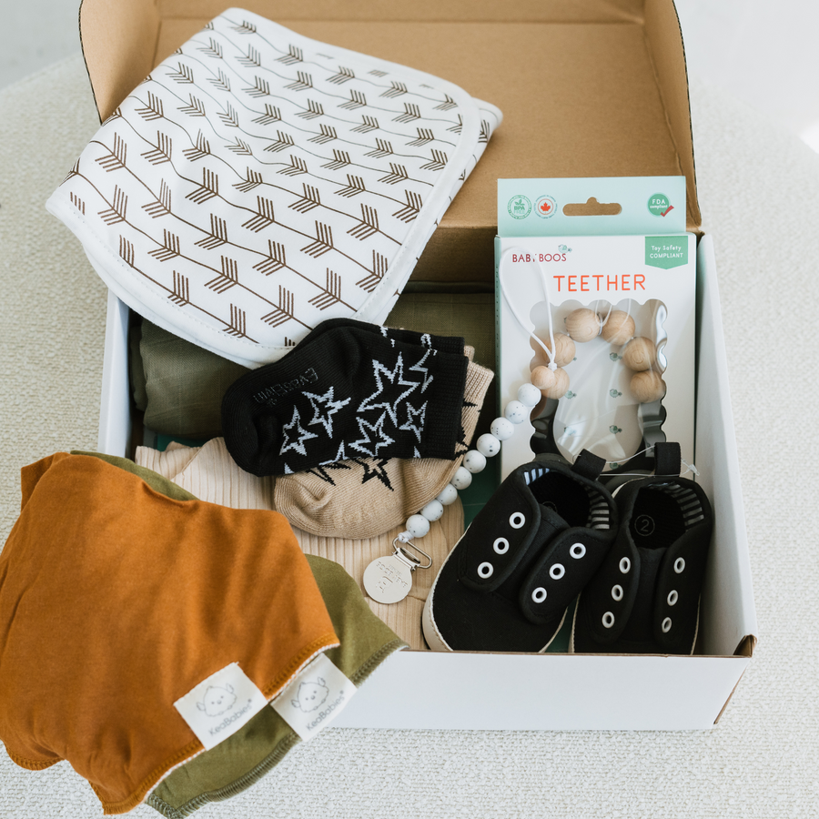 A flat lay of a gender-neutral Deluxe-Bae Box gift set, displayed in an open white box. Includes a muslin burp cloth with a brown arrow pattern, two pairs of infant socks (one black with white stars, one tan), a neutral-toned teether, black baby shoes, a pacifier clip with wooden beads, and two soft bibs in mustard and olive green.