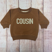 A brown organic cotton sweatshirt with the word “COUSIN” printed in bold, beige letters across the front, laid flat on a rustic wooden background.