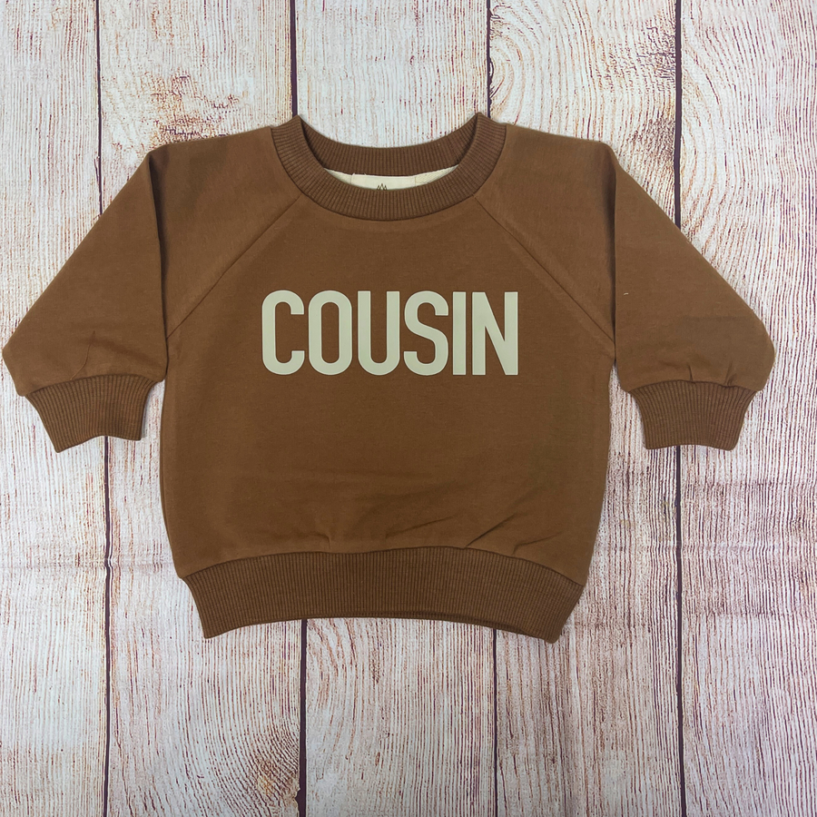 A brown organic cotton sweatshirt with the word “COUSIN” printed in bold, beige letters across the front, laid flat on a rustic wooden background.