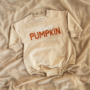 A neutral-toned baby bubble romper laid out on soft, beige fabric, featuring the words “Cutest Pumpkin” in bold, fall-inspired lettering.