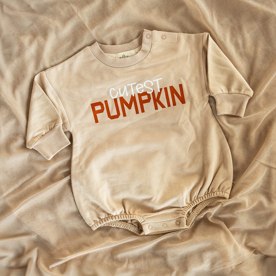 A neutral-toned baby bubble romper laid out on soft, beige fabric, featuring the words “Cutest Pumpkin” in bold, fall-inspired lettering.