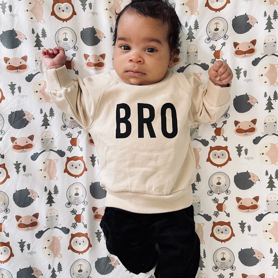 The baby is wearing a beige-colored crewneck sweatshirt with the word ‘BRO’ printed in bold black letters across the chest, paired with black pants. The baby is lying against a woodland theme blanket. 